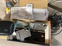 Top view of two Casio Exilim digital cameras in original box along with USB cradle chargers, cords, manuals, and a small carrying case.