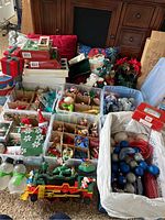Multiple containers and trays with assorted Christmas ornaments, pillows, poinsettia, stockings, and boxes displayed on floor and furniture.