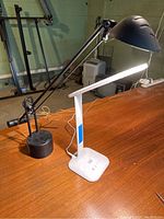 Black adjustable arm desk lamp on wooden desk, showing arm, round base and dome-shaped shade.