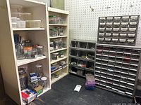 Photo showing three white shelving units filled with various jars and containers of fasteners and hardware items alongside a large multi-drawer organizer with labeled drawers