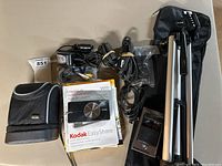 Full lot including Kodak EasyShare V610 camera case, user guides, tripod in carrying bag, and assorted cables and chargers laid out on a table.