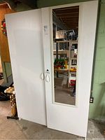 Front view of large white laminate storage cabinet with two doors, one mirrored, in basement setting.