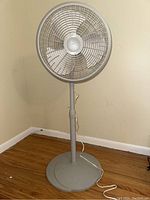 Front view of Lasko adjustable height floor fan showing the round fan cage, blades, and part of grey stand with white central hub.