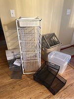 Photo of white metal frame with five wire baskets, plastic basket on top, gray plastic bins and black mesh trays on floor.
