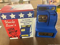 Photo of the Uncle Sam's 3-Coin Register Bank next to its original box showing front and side angle