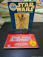 Photo showing the Star Wars jigsaw puzzle box with Tusken Raider image and Star Wars blueprints pouch with red cover on a table.