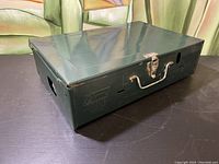 Green metal rectangular carrying case for Coleman camp stove, latch closed, showing surface wear.