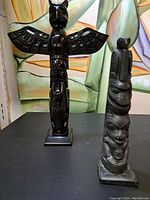 Front view of two Haida-style cast resin totem sculptures on a black surface with a colorful abstract background.