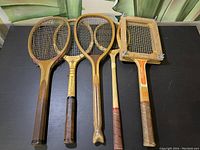 Lot of five vintage wooden tennis rackets with different head shapes and stringing conditions laid on black surface under green drapery