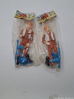Photo of two vintage Cowboy Set action figures in original clear plastic bags with paper header cards showing packaging condition and accessories.
