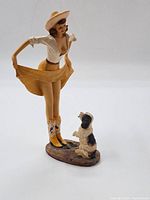 Front and side view of the porcelain figurine showing woman with hat, blouse, tan skirt and boots, and a small dog sitting at her side.