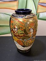 Front view of the vase showing hand-painted figures in traditional Japanese attire with gold halos and robes, detailed gold borders and patterns.