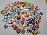 A wide shot displaying a large assortment of vintage buttons and pins scattered on a white surface. Various colors and printed slogans are visible.