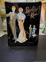 Front view of the black vinyl Barbie and Ken carry case, showing vintage graphics of Barbie and Ken dressed in formal attire with gold lettering.