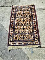 Full view of vintage hand-woven wool carpet laid on concrete, showing overall design and condition with some frayed edges and wear.