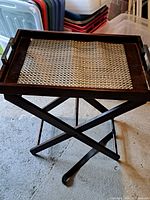 Full view of vintage wooden folding tray table standing on a concrete floor surrounded by storage bins. Shows its X-shaped folding legs and woven center panel.