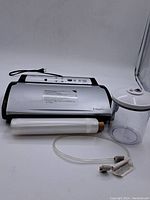 FoodSaver vacuum sealing machine with vacuum sealing bag roll, clear plastic container with lid, and vacuum hose accessory arranged on white surface.