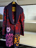 Full view of women's knitwear jacket with large collar and vibrant floral patterns in multiple colors hanging on a wooden hanger.