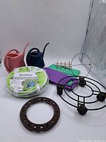 Photo showing two plastic watering cans (pink and dark blue), SunMate collapsible gardening bag in packaging, purple and green foam kneeling pads, bamboo plant support ladder, and two circular metal plant supports.