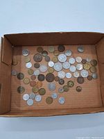 Top view of assorted world coins in cardboard box showing multiple small to medium size coins in various metals and colors.