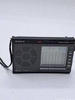 Front view of the Sony portable radio showing speaker, display, buttons, and wrist strap.