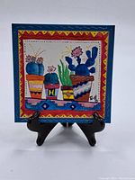 Full front photo of the colorful handcrafted ceramic tile with potted cacti and geometric border pattern on a black stand.