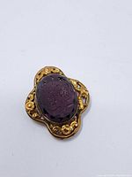 Front view of the carved glass lapel centerpiece set in an ornate gold-tone metal frame, showing the floral carving on purple glass.