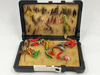 Open plastic case displaying a large assortment of vintage fly fishing jigs attached to the upper foam insert, and various brightly colored fishing lures placed on the lower foam insert.