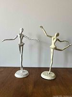 Front view of two white abstract ballerina figurines on wooden surface, positioned side by side against a plain wall background.