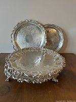 Four silver plated trays of different sizes with engraved designs and ornate edges arranged on a wooden surface