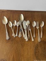 Photo showing a lineup of assorted silverware pieces including spoons, forks, and serving utensils arranged on a wooden surface.