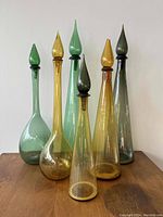 Six tall slender blown glass bottles with pointed stoppers in green and amber shades on wooden surface against plain white background.