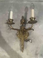 Image of a brass electric wall fixture with two candle-style light bulbs, ornate decorative arms and base, measuring approximately 17" tall by 10" wide as provided by seller.
