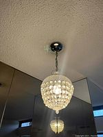 Photo of a ceiling-mounted chandelier with a clear crystal bead basket shade, hanging from a metal chain attached to a chrome ceiling fixture. The light is on, showing sparkling reflections.