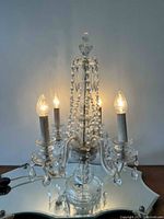 Crystal chandelier lamp with electric bulbs lit, placed on a mirrored decorative tray on wood surface against a plain wall background.