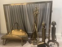 Photo of brass fireplace set including firewood holder, tool set in stand, metal mesh screen, and pair of metal andirons.