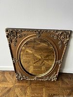 Framed mirror leaning against white wall, showing decorative brown frame with floral pattern and circular mirror center.