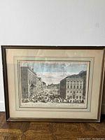 Framed vintage print depicting a European city street market with people and carriages between large classical buildings, showing sepia/black and white tones with double matting in a wooden frame.