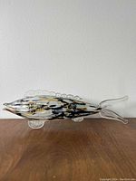Glass blown fish sculpture on wooden surface with white wall background, showing full side profile and detailed fins and pattern