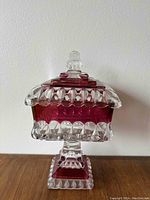Front view of a ruby red and clear decorative glass bucket with a lid, featuring intricate diamond and scalloped glass patterns and a stepped square base.
