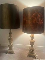 Photo of two table lamps side by side on the floor against a white wall, both with dark lampshades and beige ornamental bases, showing approximate height and design.