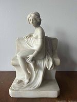 White marble statue of a seated woman against a plain wall background.