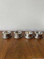Four silver napkin holders aligned in a row on a wooden surface against a plain wall, showing their size, shape and condition.
