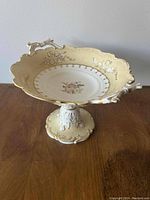 Photo of cream-colored porcelain pedestal serving dish with floral center and scalloped edges on wooden table.