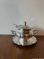 Front view of ornate silver plated gravy boat with lid on matching scalloped underplate with ladle resting