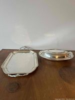 Photo of two silver plated trays placed on wooden surface, showing top view of rectangular tray and bottom view of oval tray
