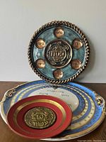 Photo shows four decorative plates stacked or arranged, highlighting the largest bronze-tone plate with Hebrew inscriptions and smaller plates in blue, white, and red with varying designs and Hebrew text.