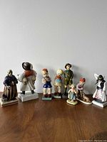 Photo of eight porcelain figurines posed on a wooden surface against a white wall. The figurines depict children and adults in traditional and classic clothing styles.