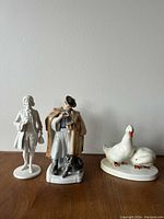 Photo shows three porcelain figurines: a white gloss man, a man in colored outfit with a dog, and two white geese.