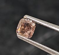 Close-up front view of cushion cut brown-pink diamond held by tweezers against dark background